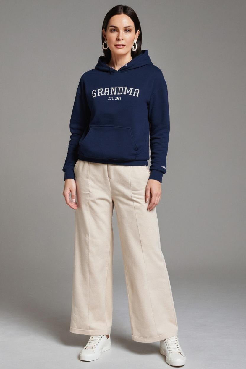 Woman wearing a navy hoodie with 'GRANDMA' text, beige pants, and white sneakers on a gray background