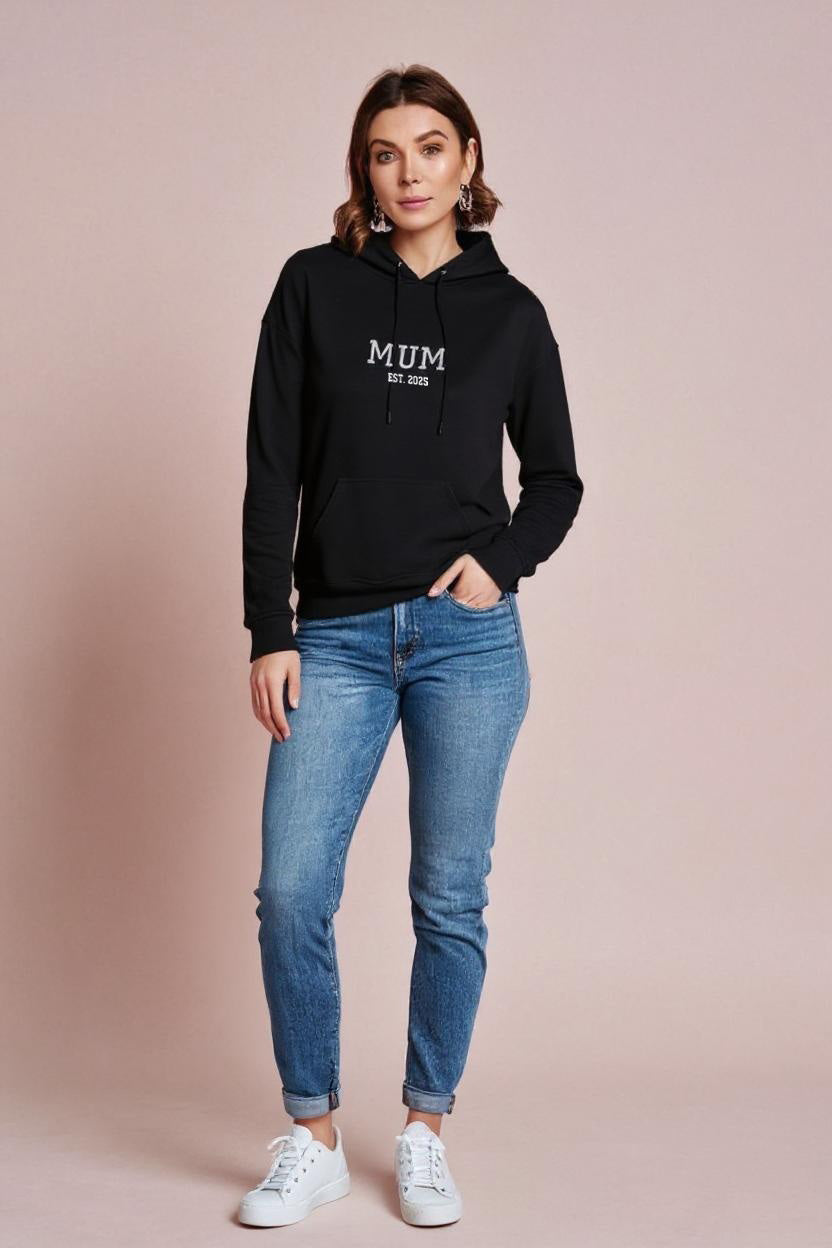 Woman wearing a black hoodie with 'MUM' text and blue jeans on a beige background