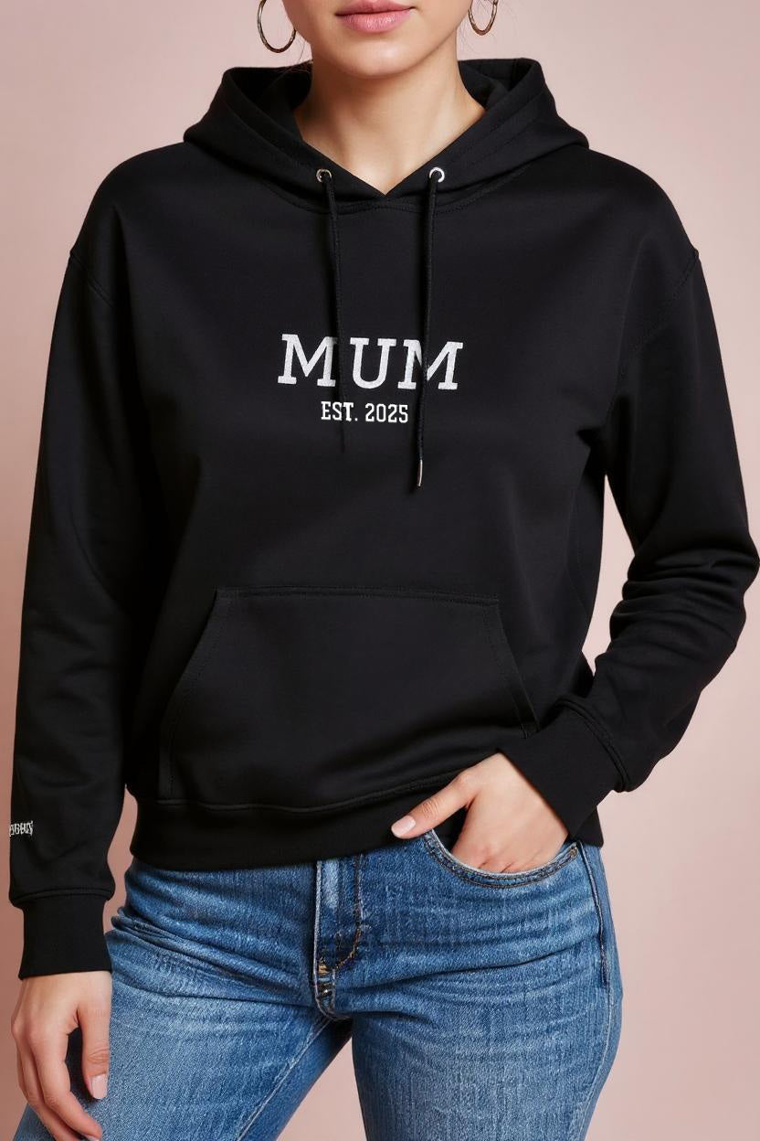 Black hoodie with 'MUM EST. 2023' text worn by a person on a pink background