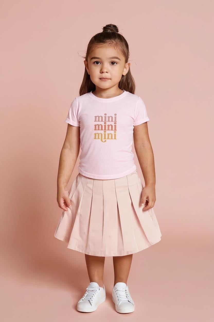Young girl wearing a pink t-shirt with 'mini' text and a light pink skirt on a pink background