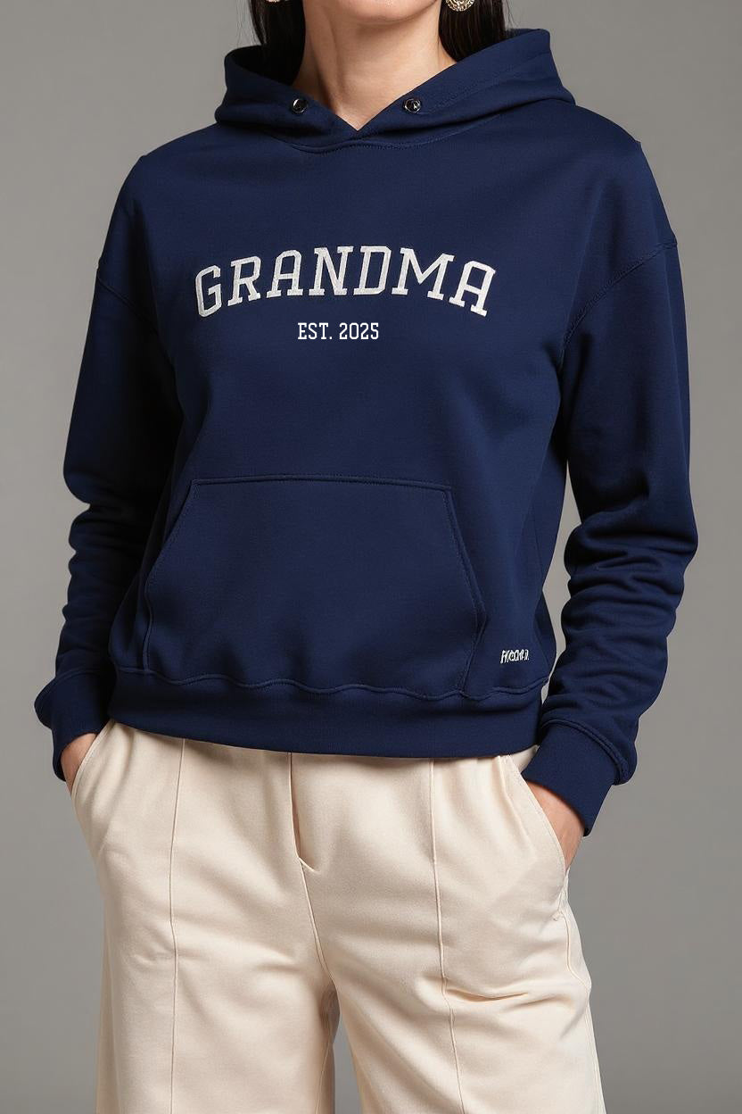 Navy blue hoodie with 'GRANDMA EST. 2025' text worn by a person on a gray background